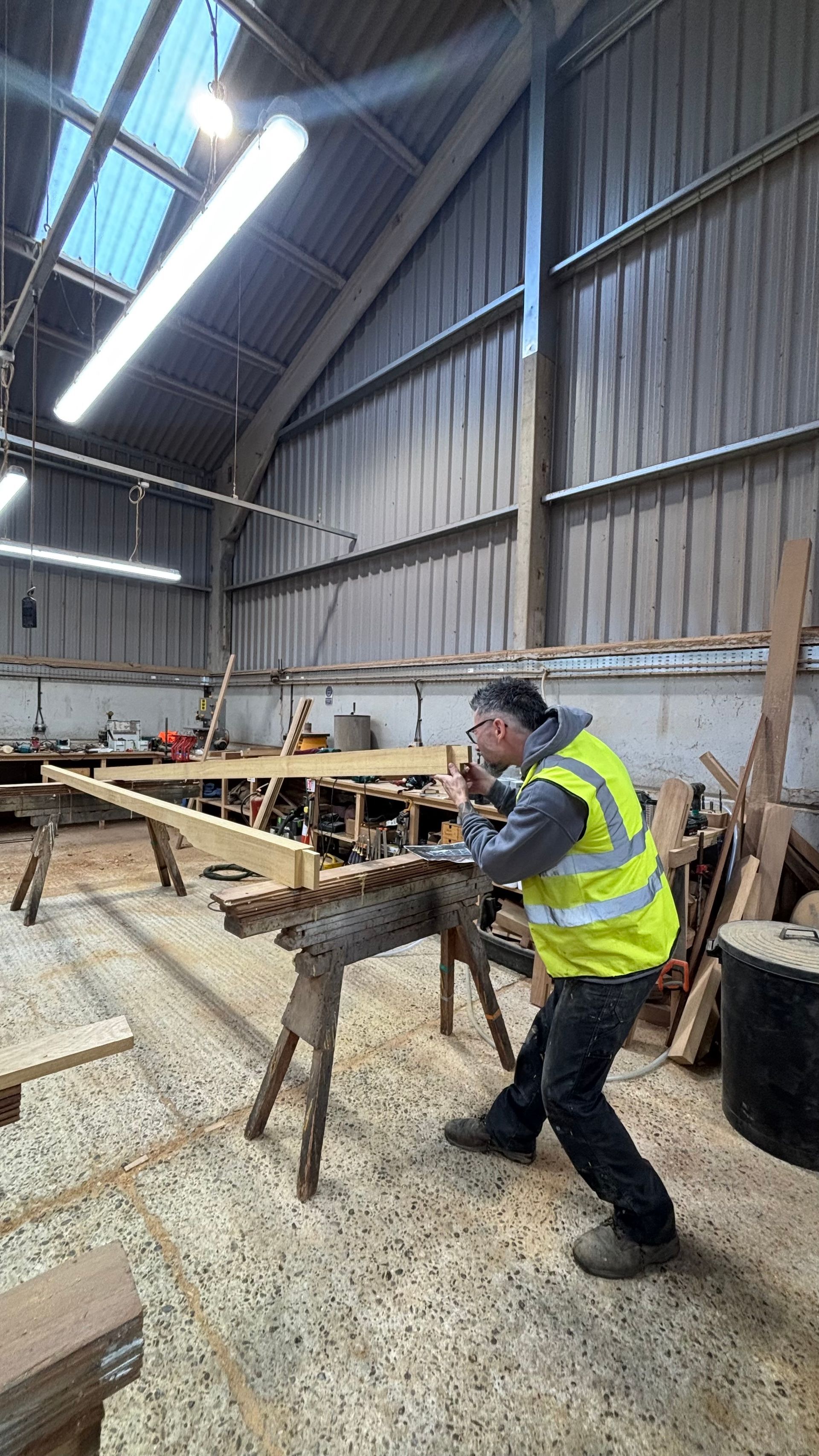 Worker in a high-vis vest carrying a sawhorse in a large workshop with timber and tools.