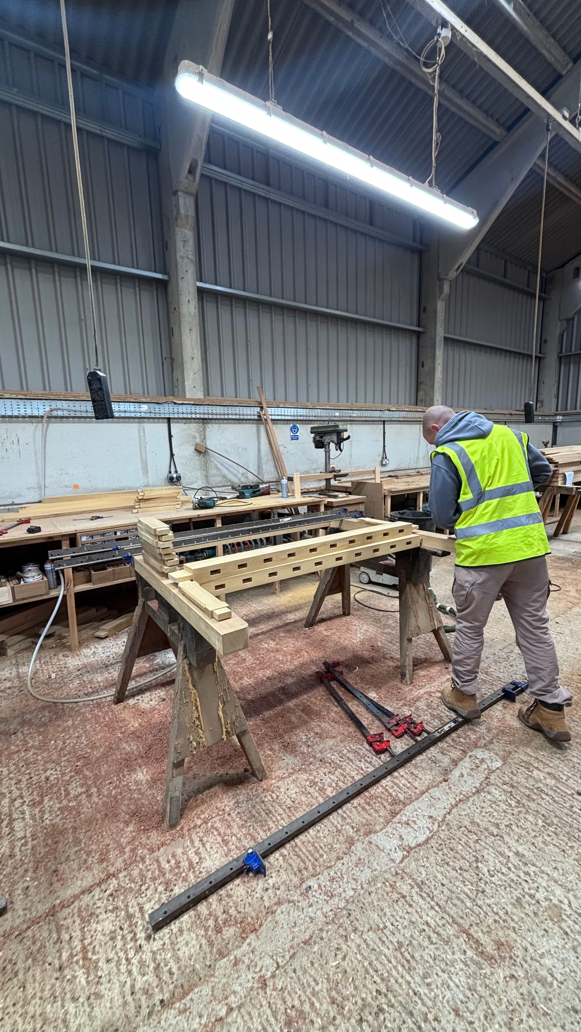 Worker in a yellow vest grinding a metal frame in an industrial workshop.