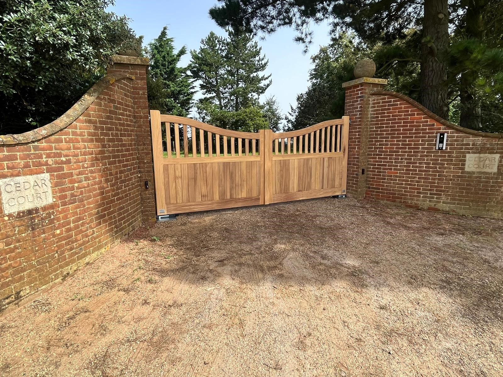 Wooden gate between brick walls at a gravel driveway entrance, surrounded by trees.