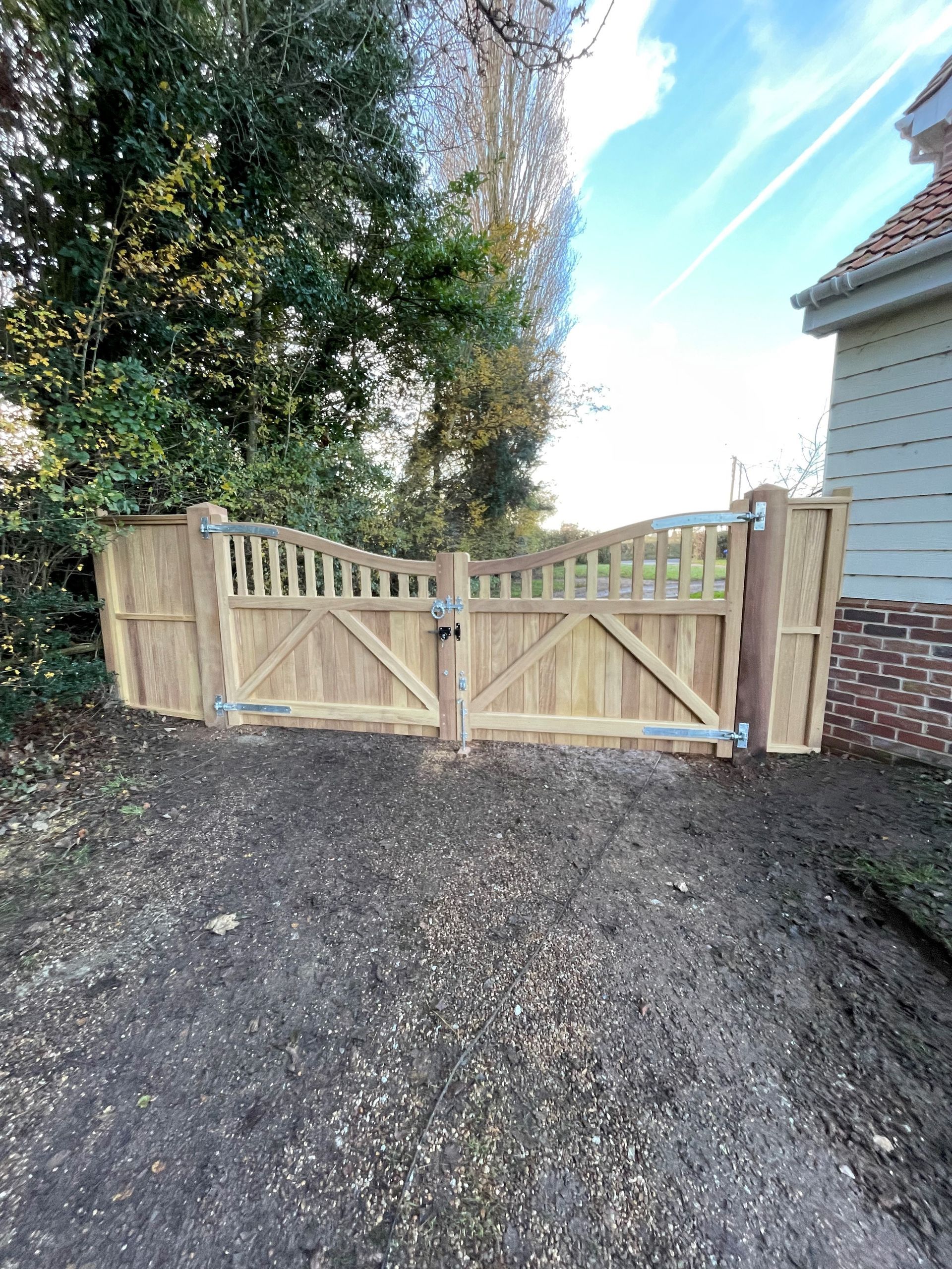 Wooden double garden gate in a gravel driveway beside a brick house and trees