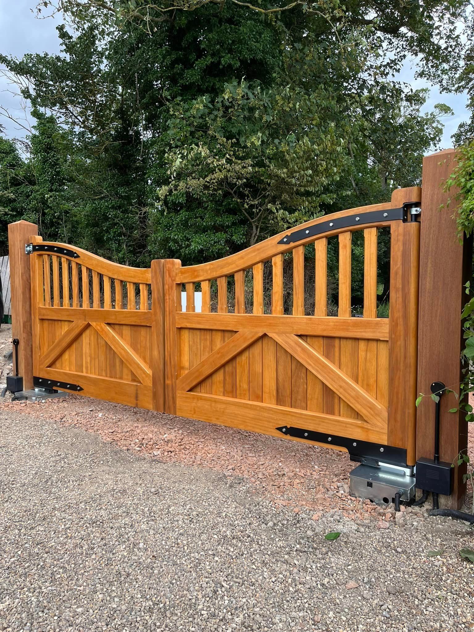 Wooden double driveway gate with black hinges on a gravel driveway, bordered by trees and shrubs.