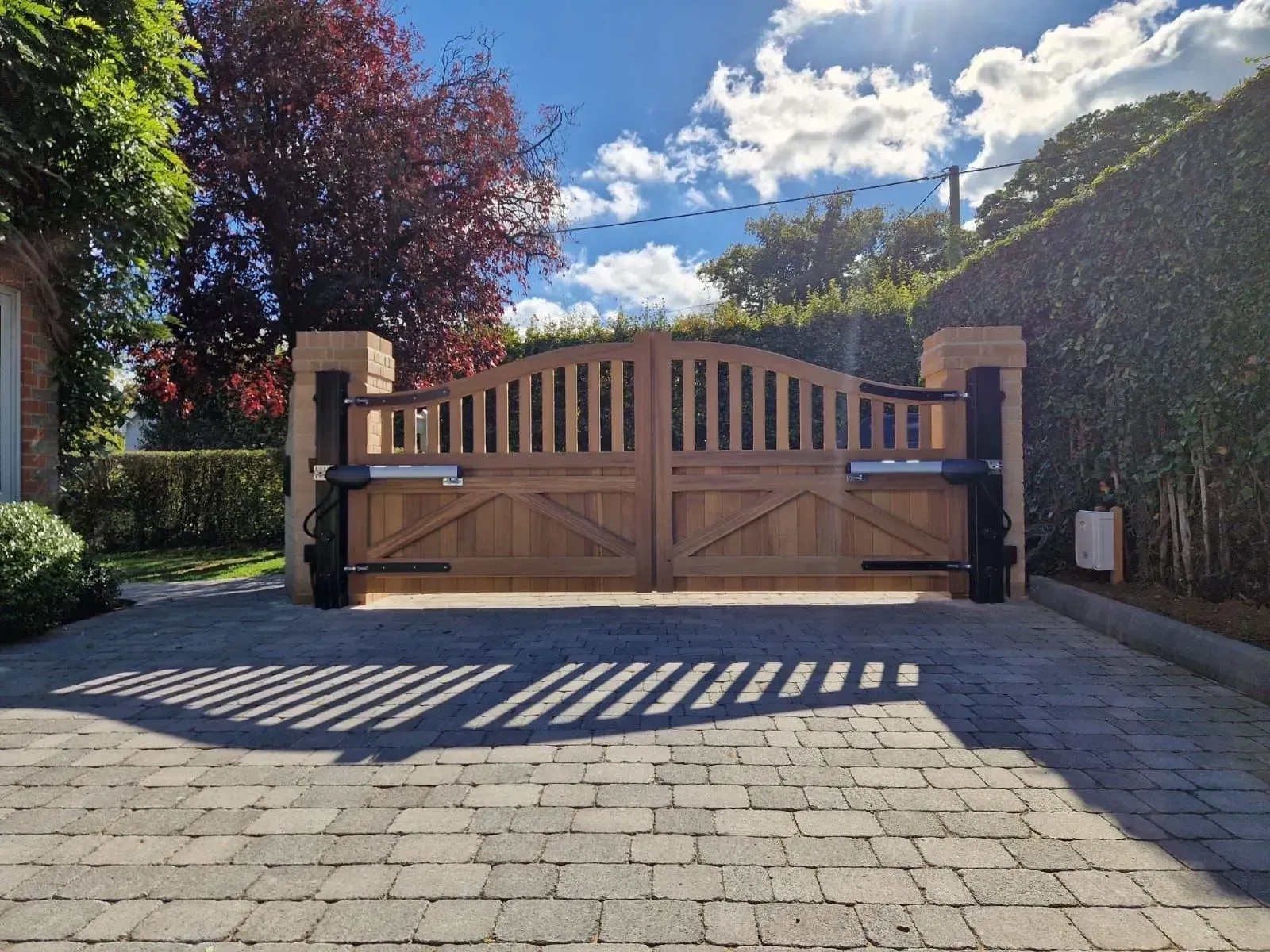 Wooden driveway gate with stone pillars and hedge, casting shadows in a sunny garden entrance