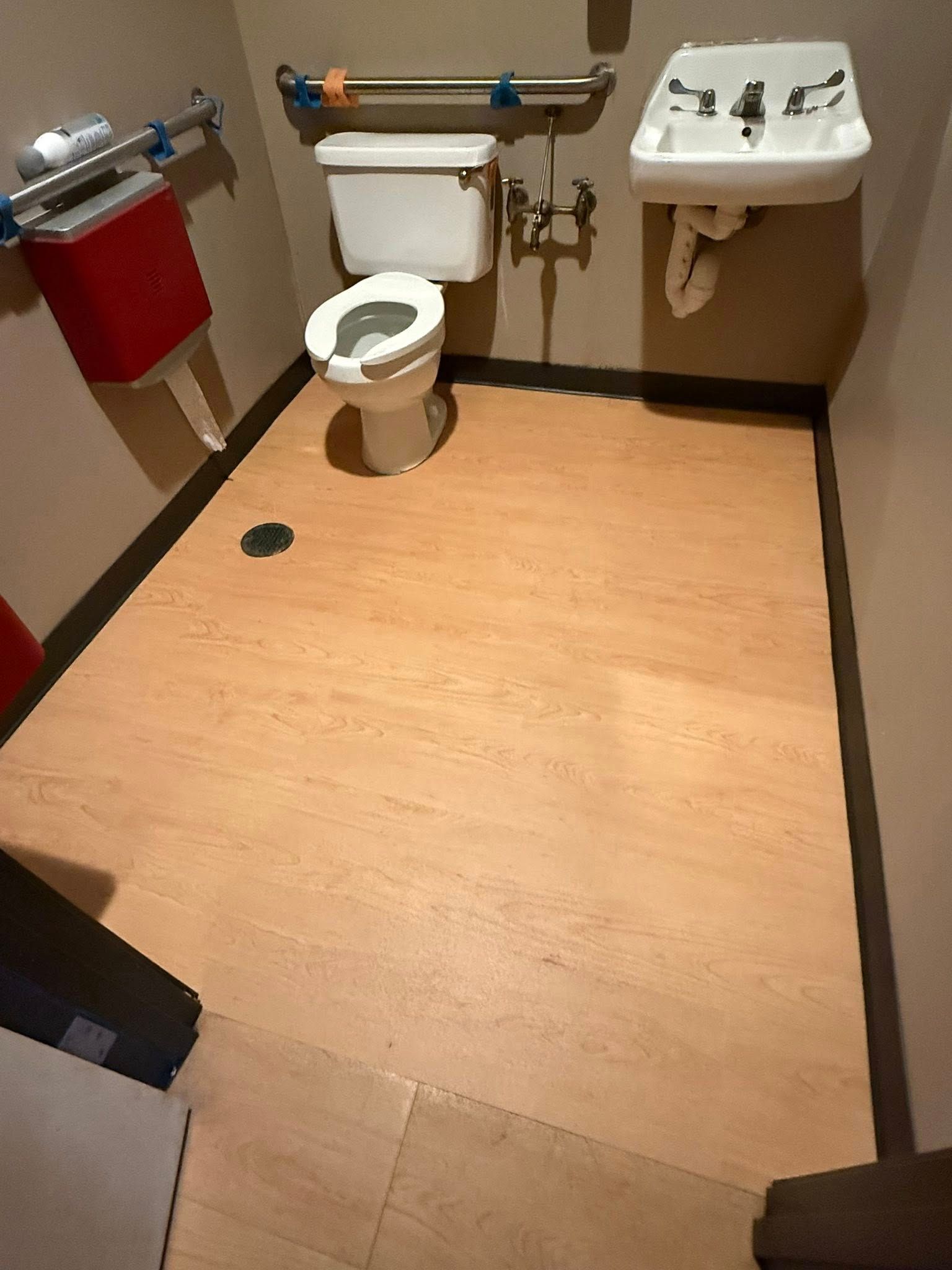 Small accessible restroom with toilet, sink, and red paper towel dispenser. Light wood-look floor, grab bars.