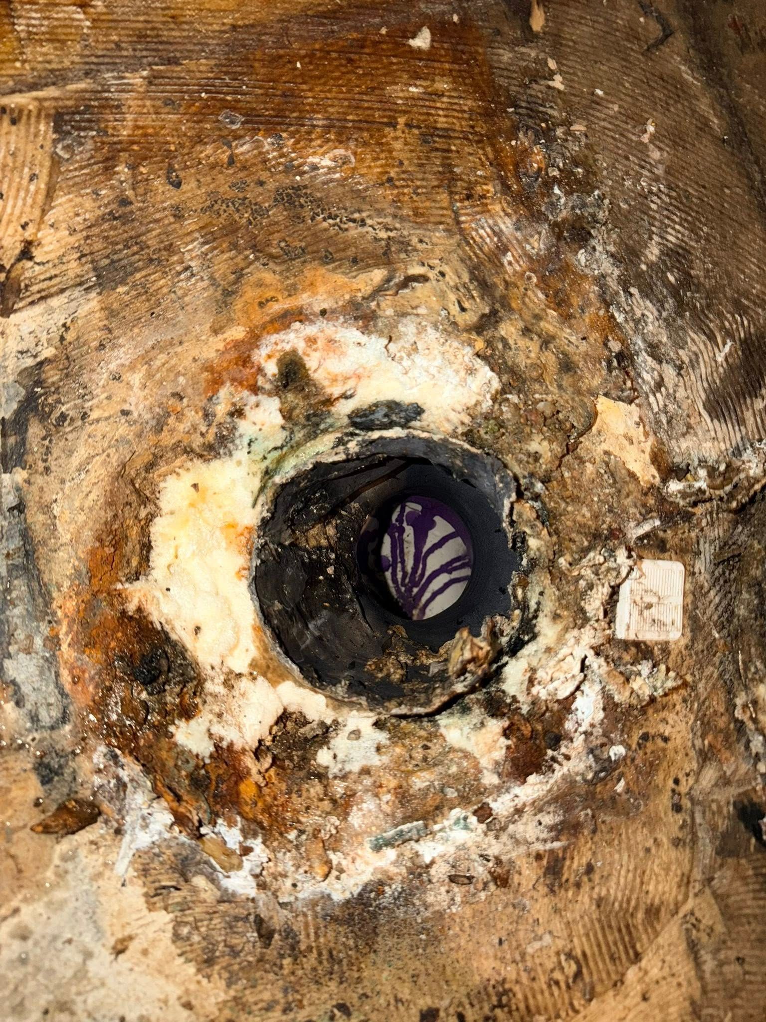 Close-up of a dark drainpipe in the center, surrounded by deteriorating light and dark brown flooring material.
