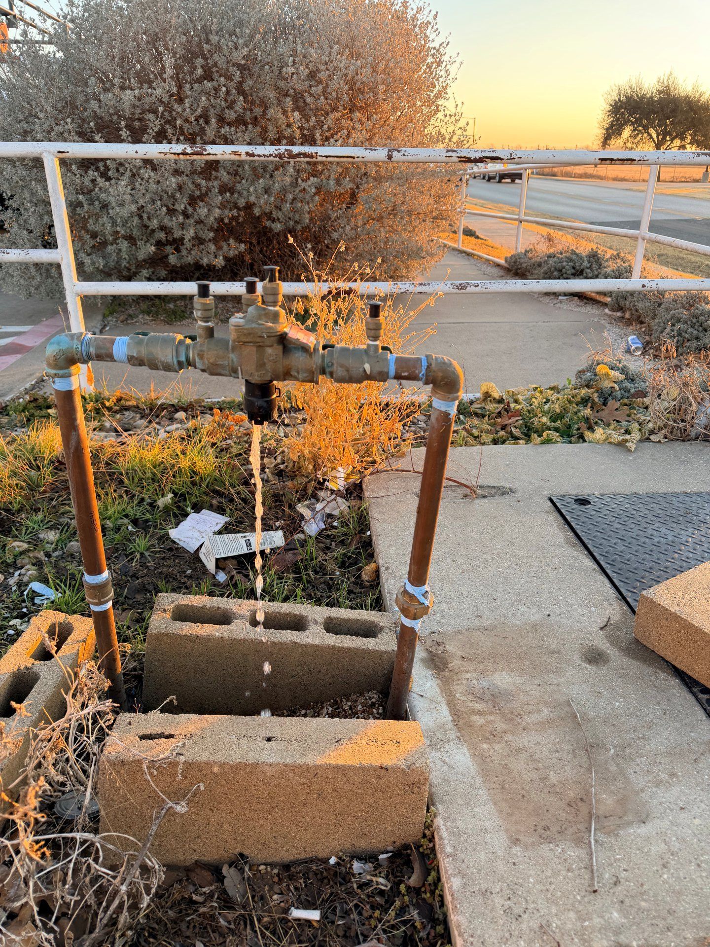 Water spraying from a broken pipe in front of a sidewalk and a white fence. The sky is golden.