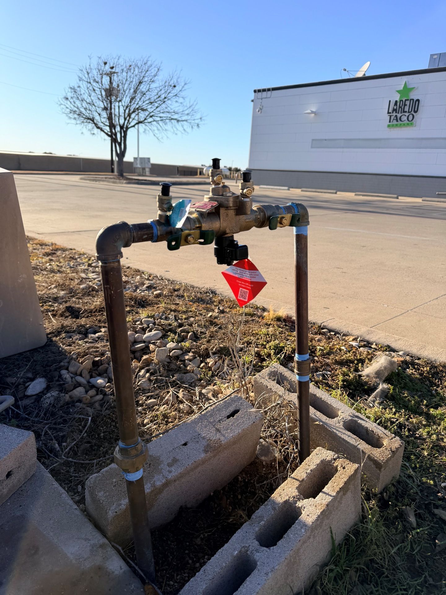 Water pipes and valves, with a red tag, secured by cinder blocks, outdoors under a clear sky.