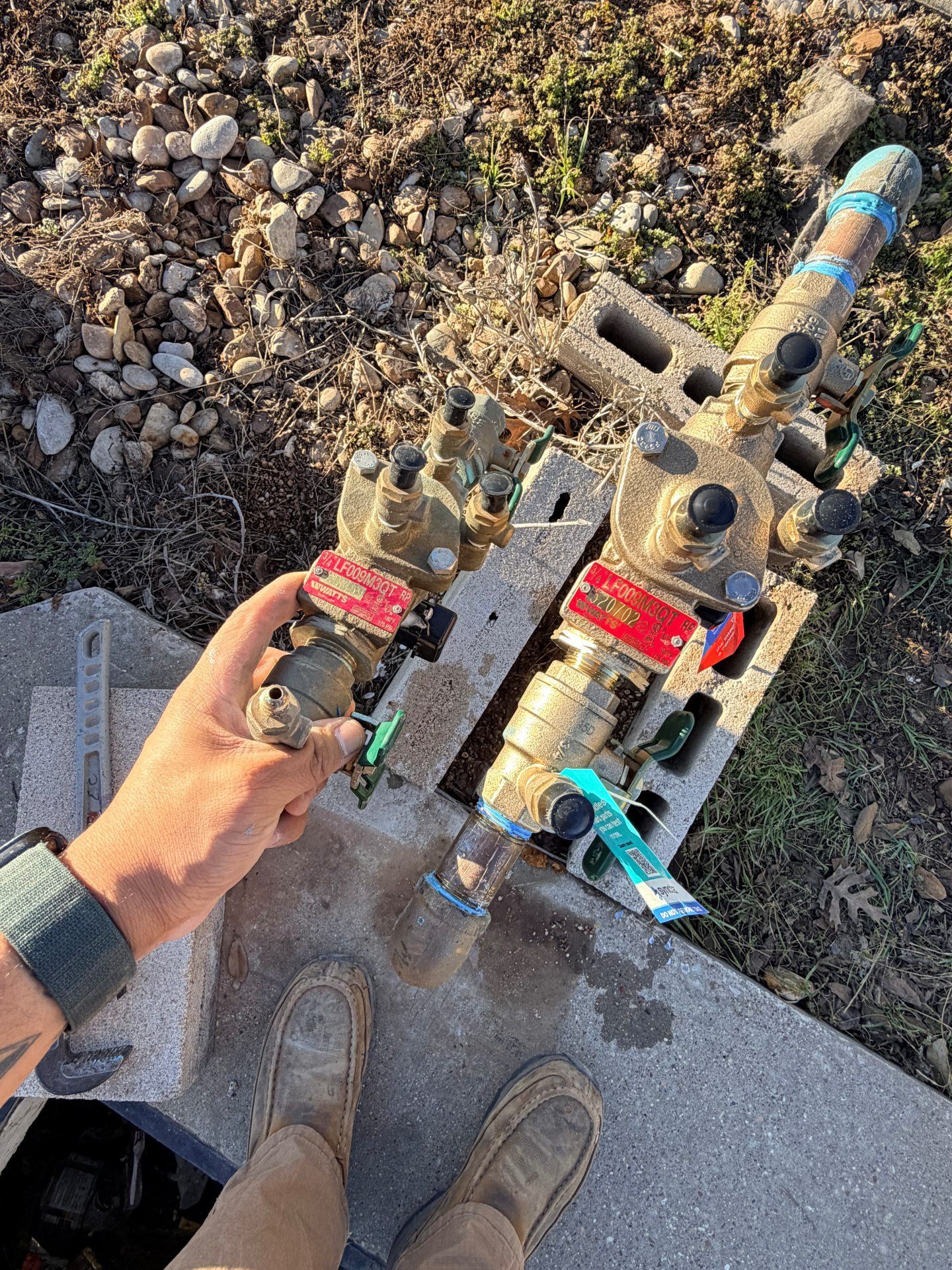 A person holds a water valve near two other valves attached to pipes and set on cinder blocks outdoors.