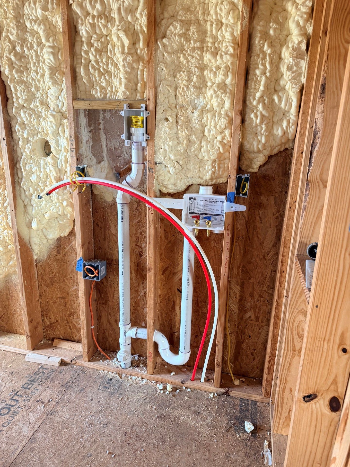 Plumbing pipes and electrical boxes installed within wooden wall studs, with spray foam insulation.