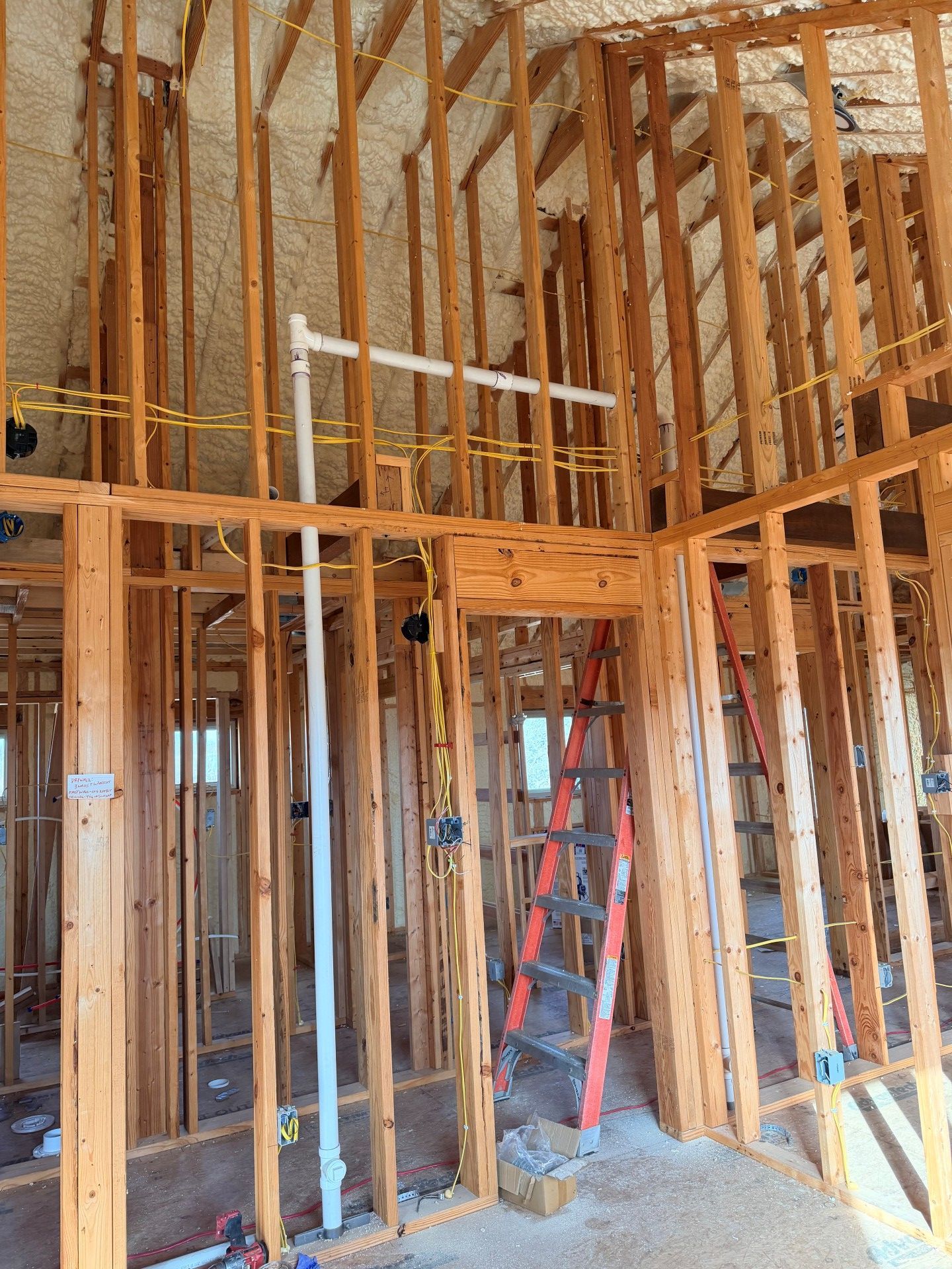 Interior view of a building under construction, wooden frame with installed plumbing and electrical wiring, ladder in a doorway.