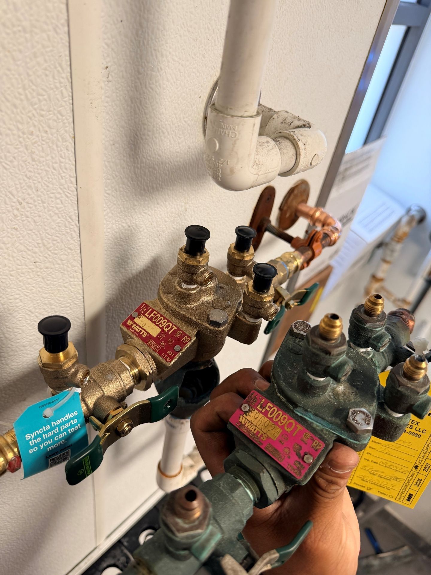 Hand holding a plumbing component near other valves and pipes on a wall.