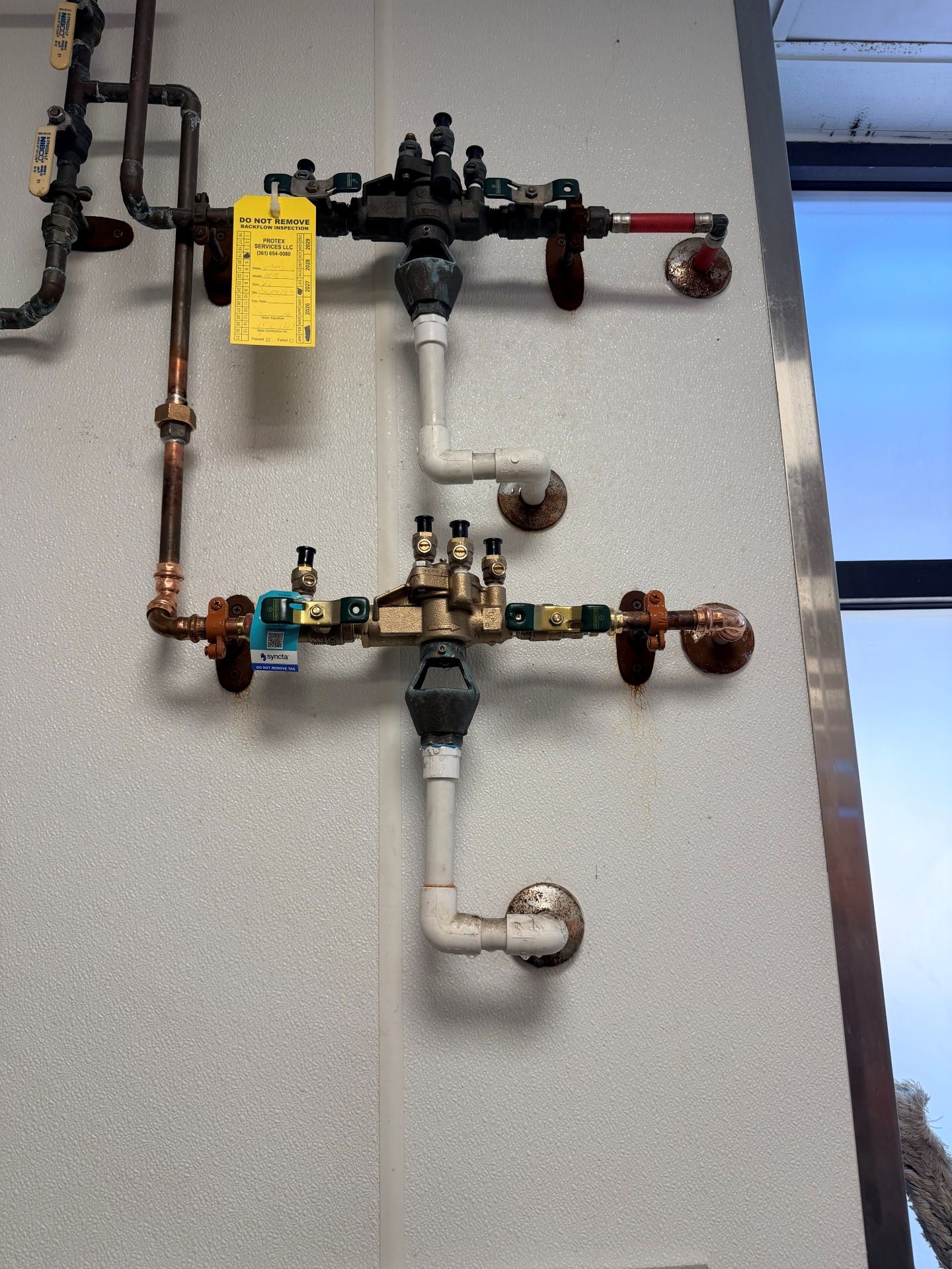 Plumbing pipes and valves mounted on a wall. Two assemblies with various fittings and a yellow tag.