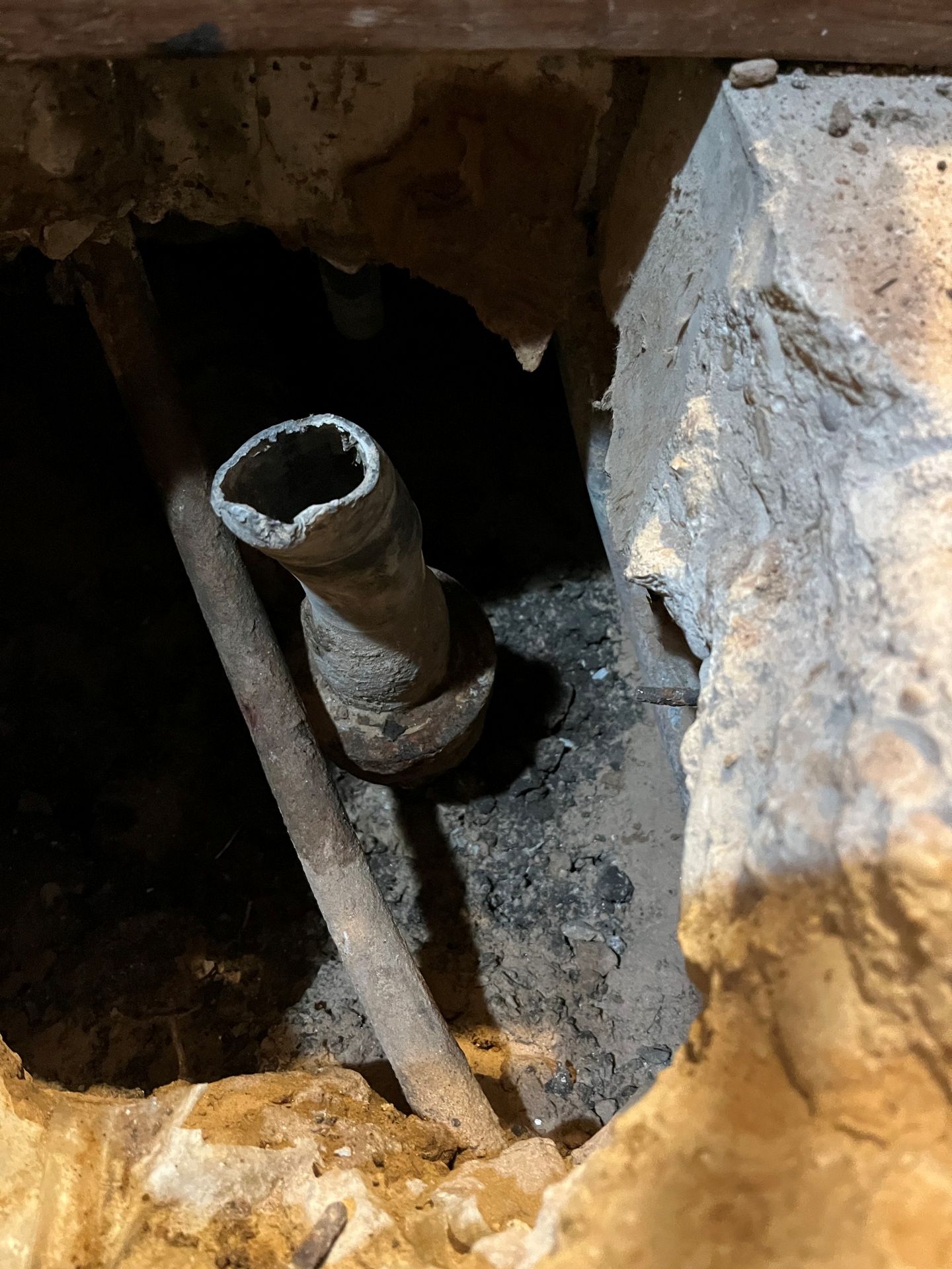 A corroded pipe elbow emerges from a dark opening in a concrete wall, revealing a damaged interior.