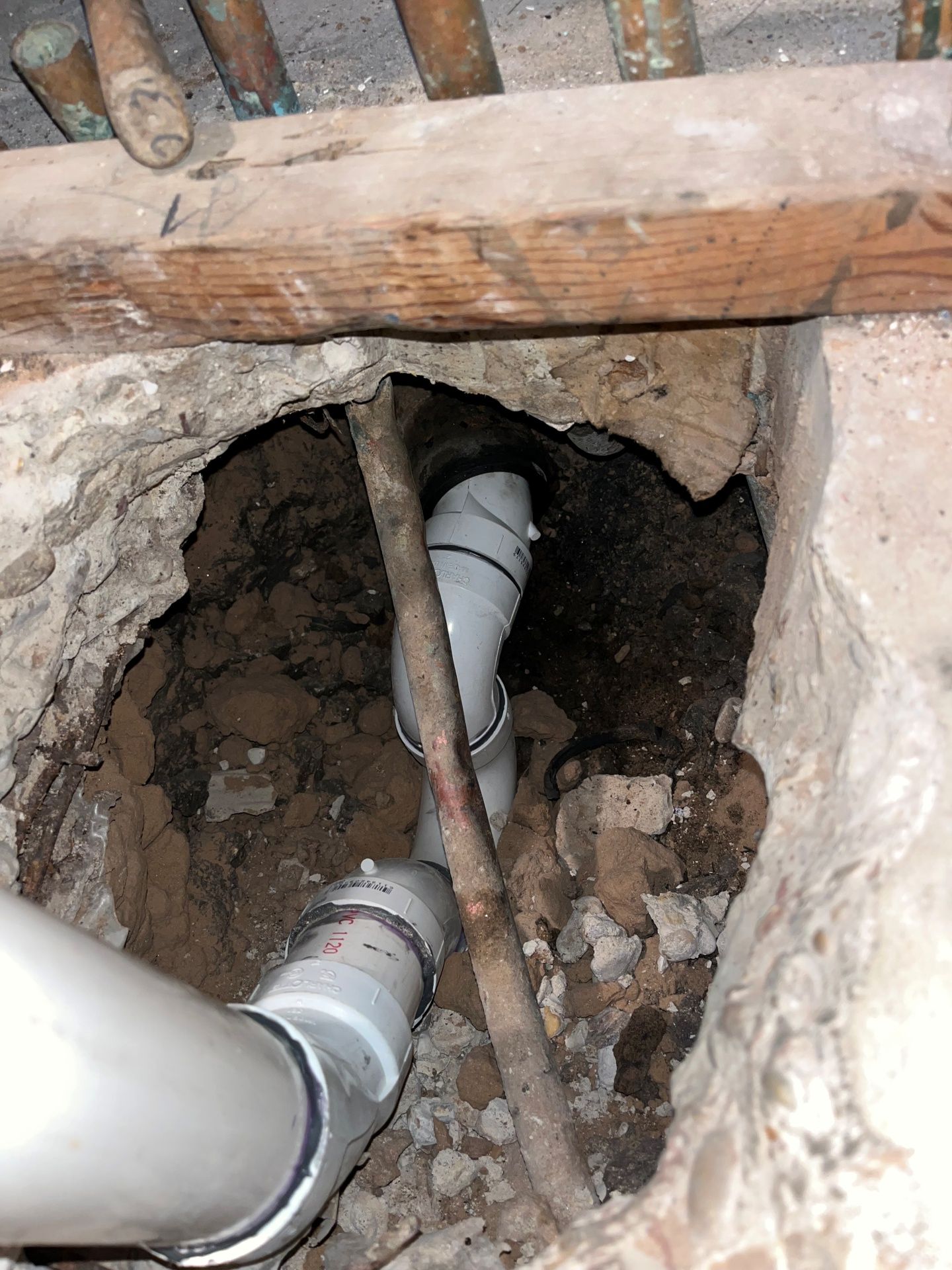 Hole in floor with white PVC pipes. A wooden beam and older copper pipes are visible above.