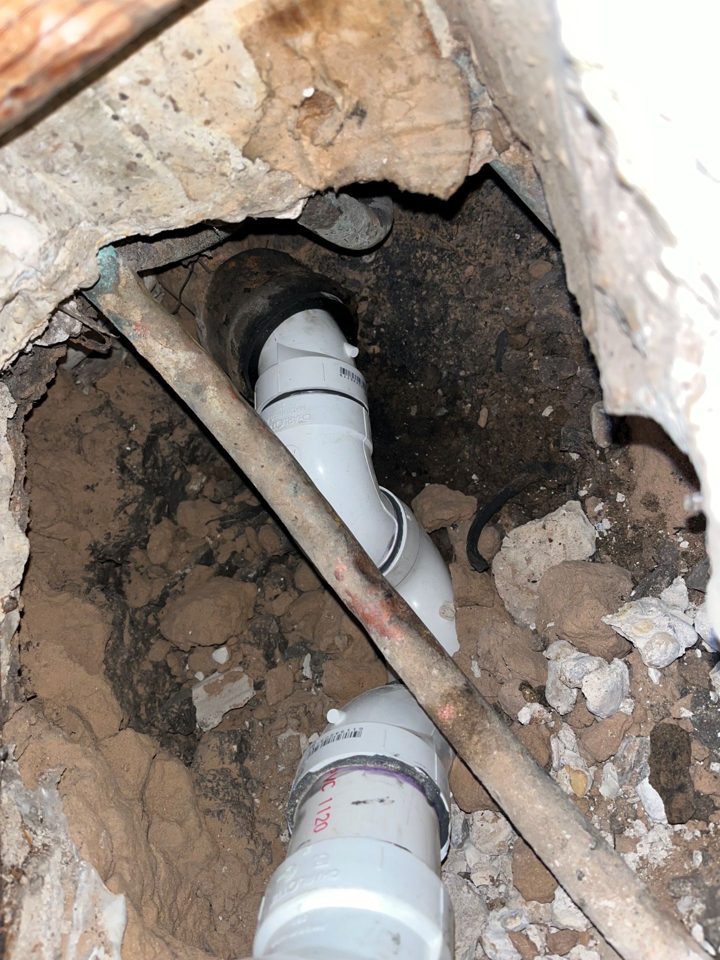 White PVC pipes and black rubber fittings in a hole in concrete. Brown wood beam spans the opening.