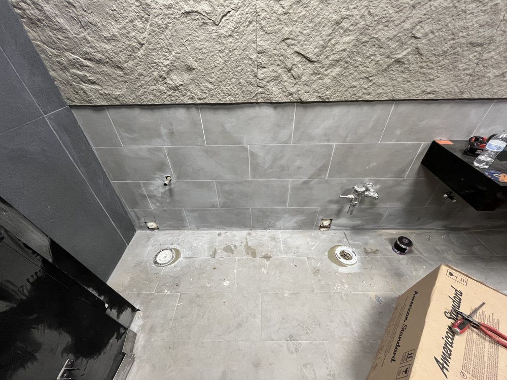 Bathroom under construction: gray tiled wall, concrete floor with toilet rough-ins, and stone-like upper wall section.