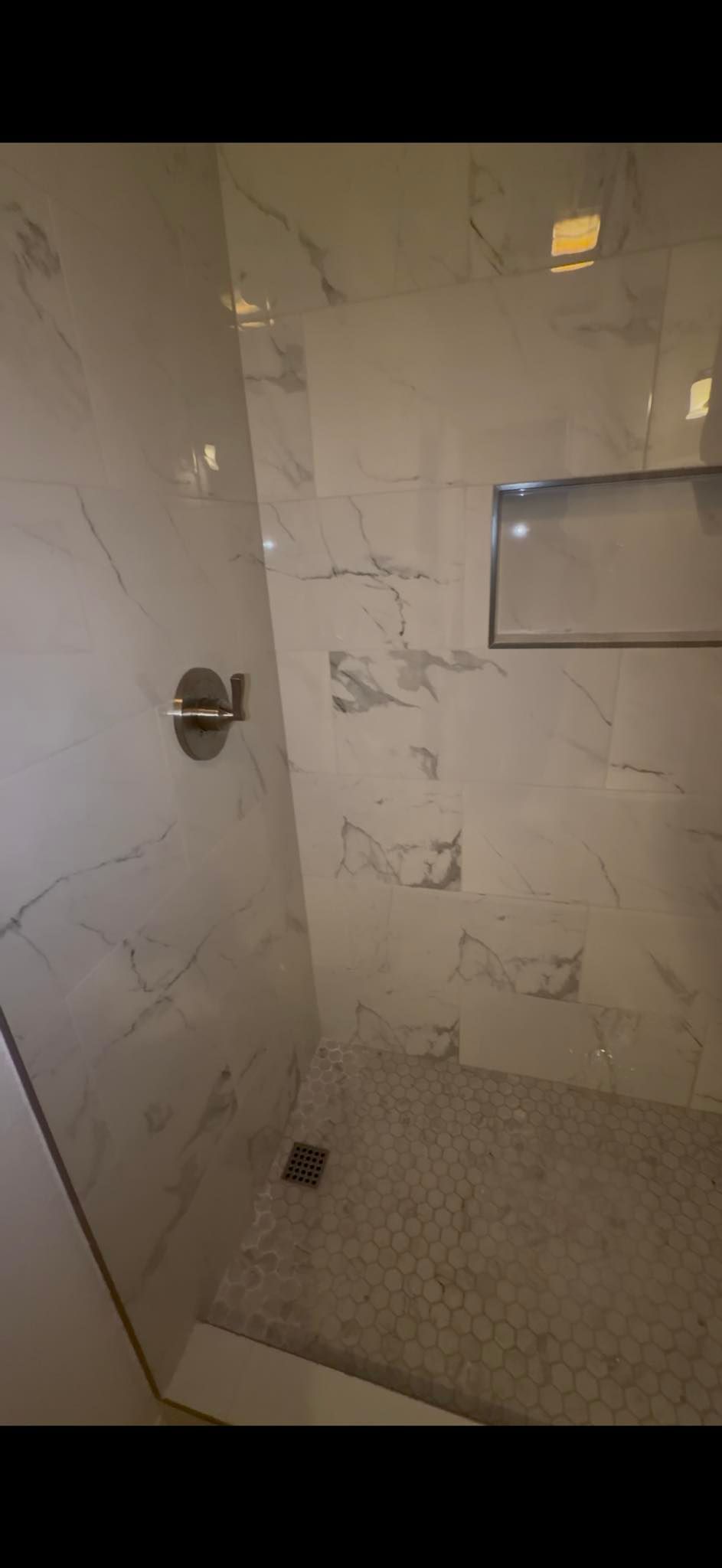 A shower with white marble tile and a built-in shelf. A silver shower handle is on the wall.