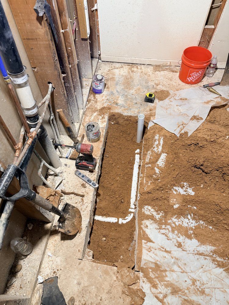 Bathroom renovation: exposed pipes in a trench, dirt, tools, and a red bucket.