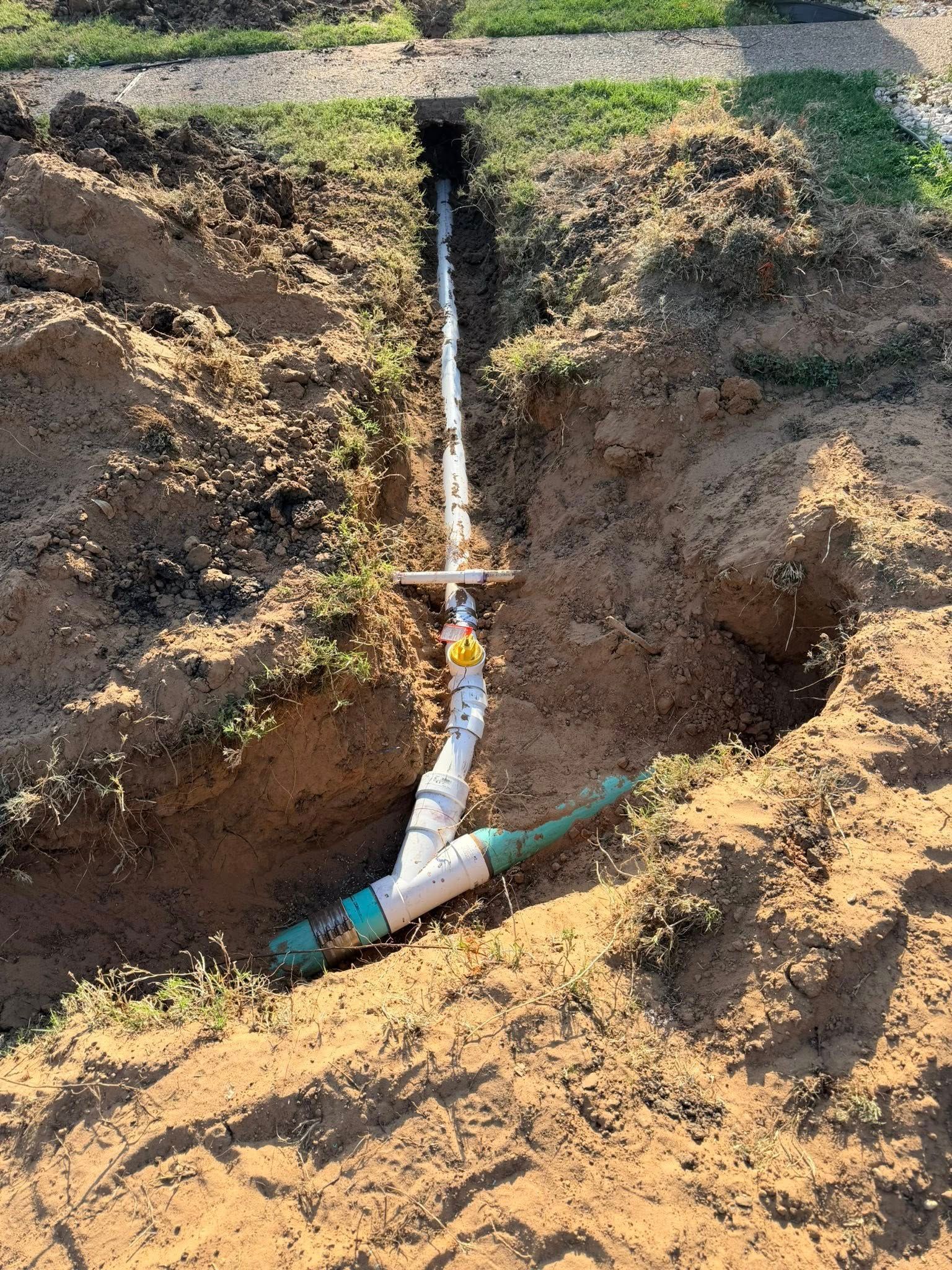 Dug trench with white PVC pipes and green sprinkler heads, installed in the ground.