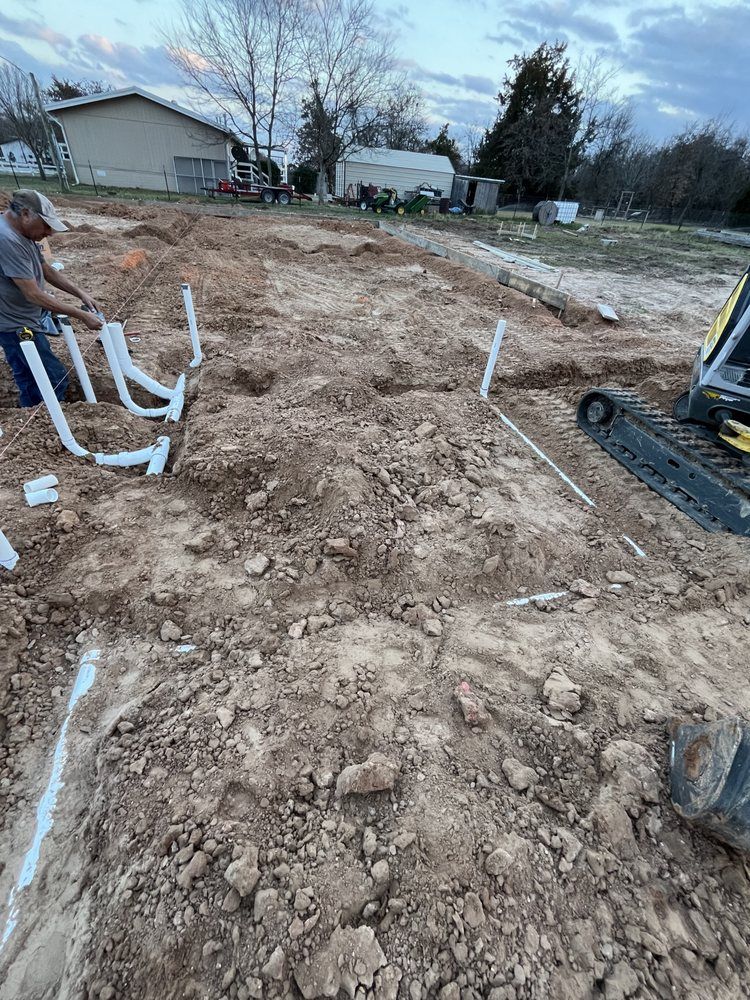 Construction site with a person installing white pipes in the dirt, next to a small excavator.