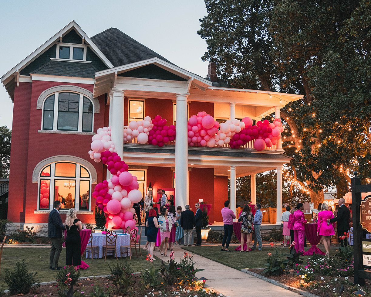A historic house decorated with pink and white balloon garlands hosts an outdoor evening party with guests.