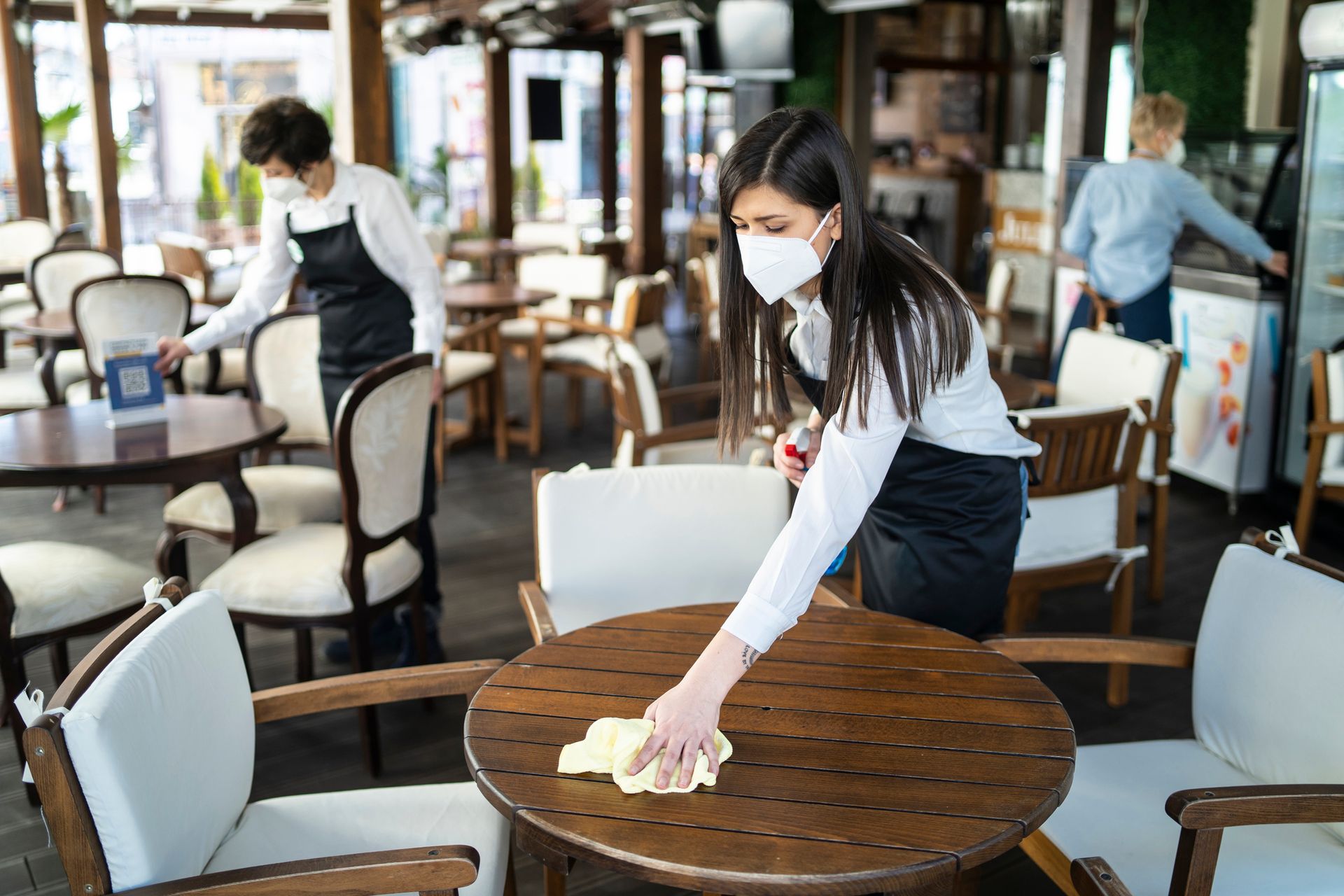 A woman wearing a mask is cleaning a table in a restaurant.