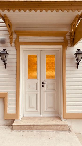 White double front door with amber glass panes beneath a tan porch awning and black wall lanterns.