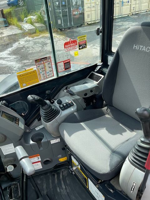 Inside a Hitachi excavator cab with joysticks, seat, and rain-speckled front window visible.