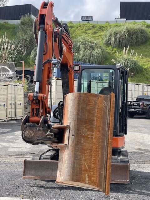 Orange excavator at a work site holding a large rusted metal sheet, with a hillside in the background