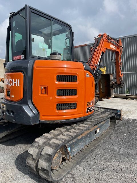 Orange Hitachi mini excavator parked on gravel lot, side view with tracked undercarriage and raised arm.