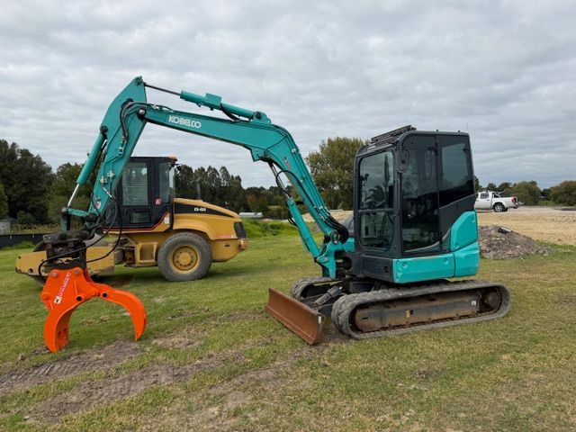 Teal excavator with orange grapple beside a yellow roller on a grassy construction site under cloudy skies