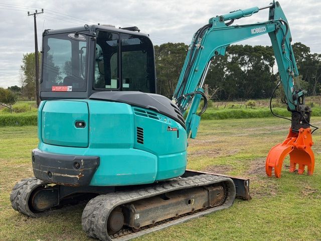 Turquoise excavator with orange grapple bucket parked on grass in an open field.