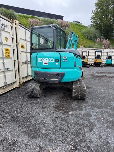 Turquoise excavator parked beside shipping containers on a gravel lot.