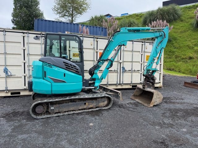 Turquoise mini excavator parked beside white shipping containers on gravel
