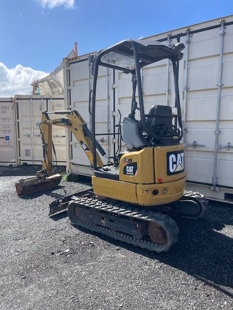 Yellow CAT mini excavator parked on gravel beside large white shipping containers