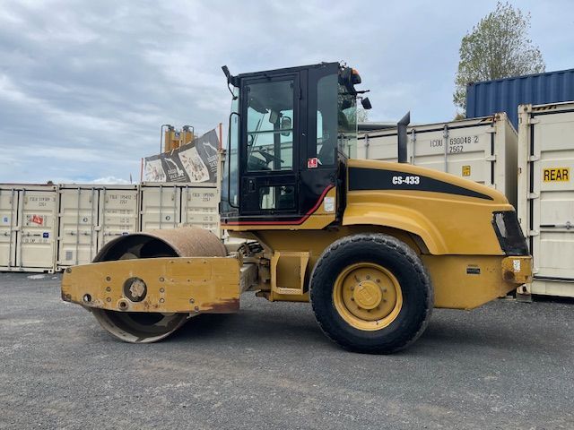 A yellow and black roller is parked in a parking lot next to shipping containers.
