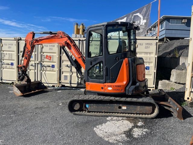 Orange mini excavator parked on gravel in front of stacked shipping containers under a blue sky
