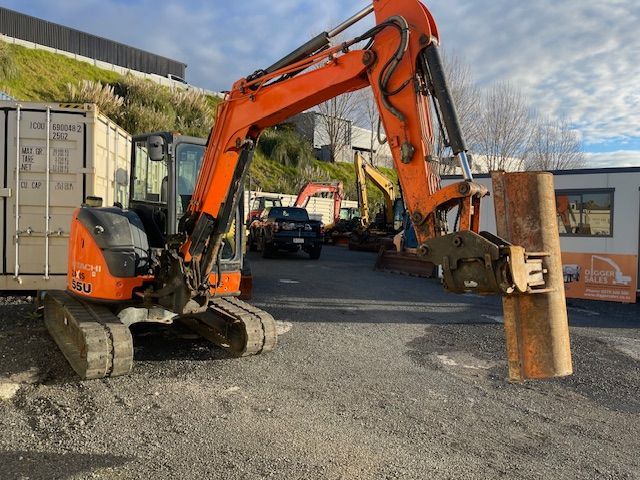 Orange excavator lifting a large metal sheet in a gravel yard with shipping containers and buildings nearby