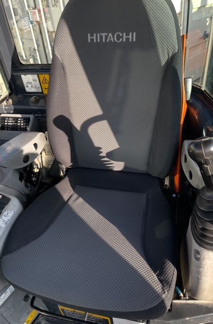 Gray Hitachi excavator seat inside a machine cab, with black armrests and a shadow across the cushion.