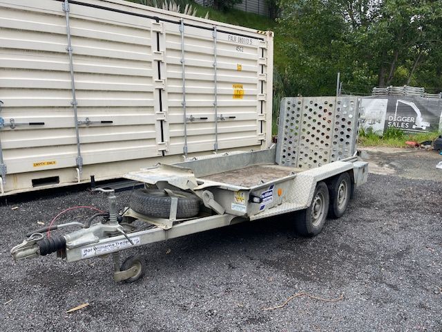 Flatbed utility trailer parked on gravel beside a shipping container, with a damaged front hitch assembly.