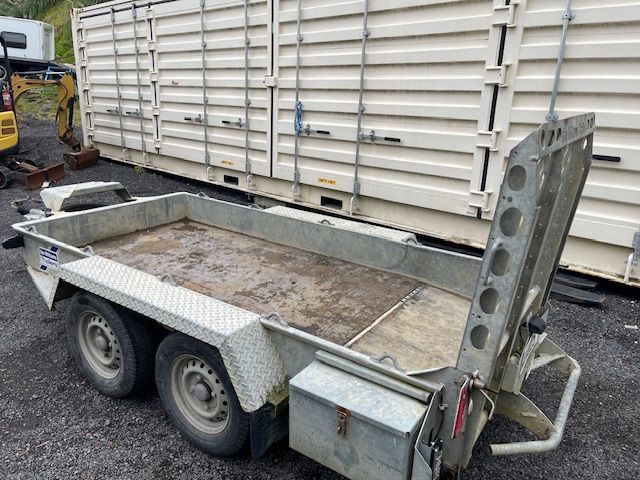 Small flatbed utility trailer with metal ramp parked beside a white truck/trailer.