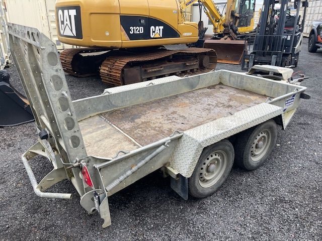 Damaged silver utility trailer with bent gate parked on gravel at a construction site, excavator behind it