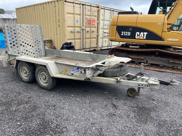 Damaged silver utility trailer parked in a gravel lot beside a yellow CAT excavator and shipping container