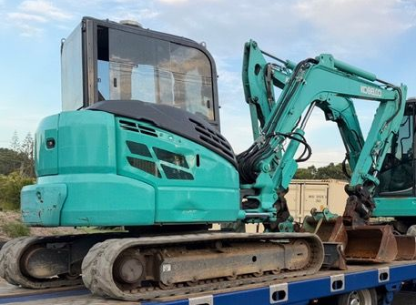 Turquoise excavator on a flatbed trailer at a construction site, with an extended arm and tracks visible