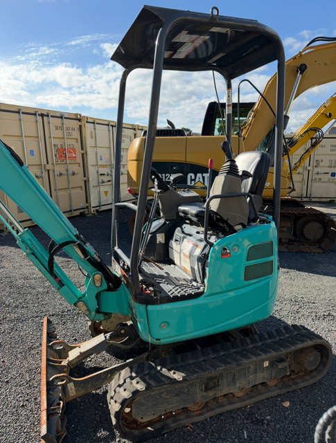 Turquoise mini excavator on tracks parked in a gravel lot with shipping containers behind it