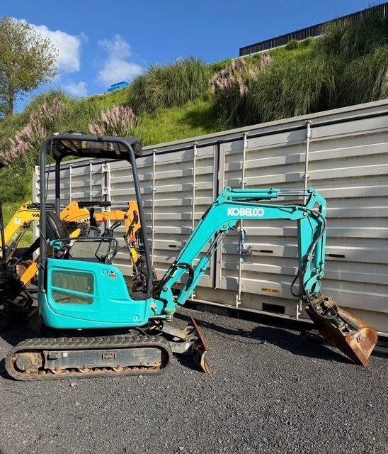 Turquoise mini excavator with bucket parked on asphalt beside a gray fence and grassy hillside.