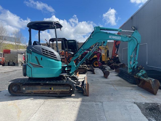 Turquoise mini excavator parked beside a warehouse on a sunny day