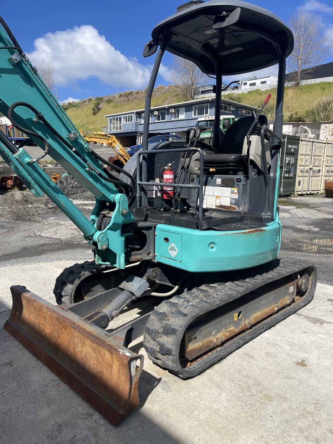 Turquoise mini excavator with front blade and canopy parked on gravel, construction site background