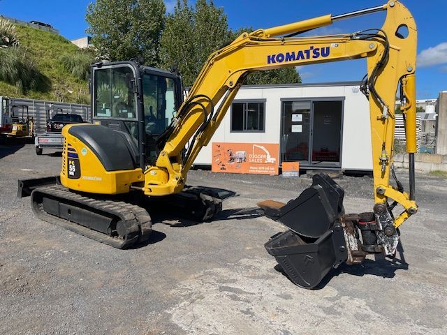Yellow Komatsu excavator with bucket attachment parked outside a small building on a gravel lot.