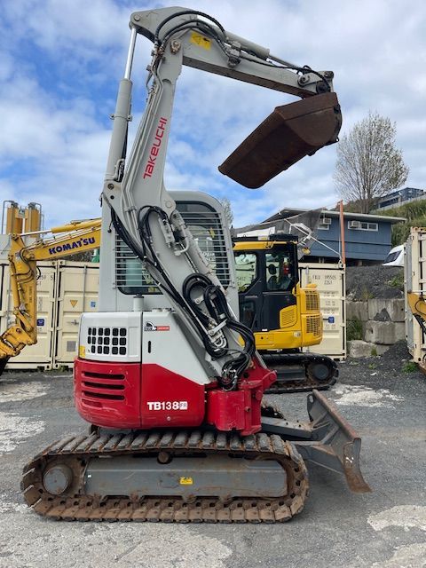 Red mini excavator with raised bucket on tracks at an outdoor construction yard