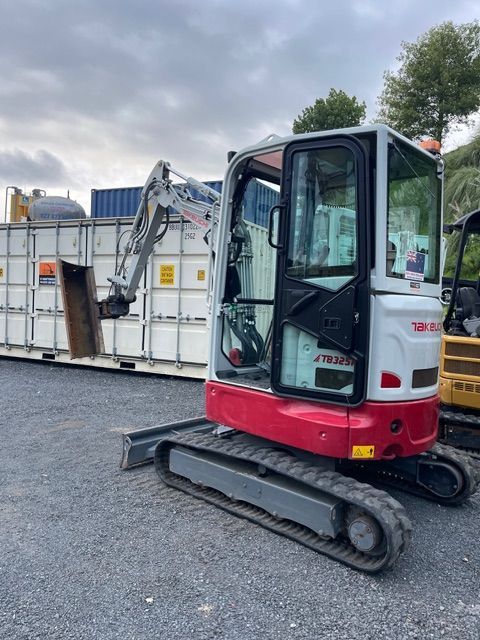 Red tracked mini excavator parked on gravel beside large white containers under a cloudy sky